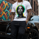 Man wearing a white t-shirt with a colorful graphic design, standing in front of a black car with graffiti in the background.